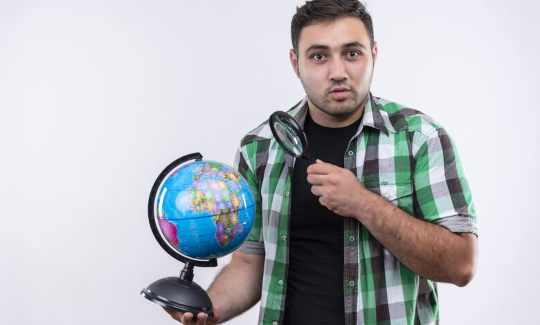 young traveler man checked shirt holding globe magnifying glass looking surprised standing white wall